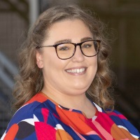 Smiling person with shoulder length blond hair wearing glasses and a colorful geometric blouse