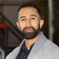 Smiling person with short dark hair and a full beard and mustache wearing a gray cardigan sweater over a black polo