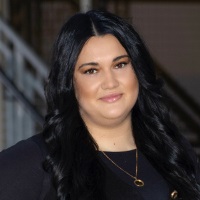 Person with long dark hair parted in the middle wearing a black blazer over a dark shirt and a gold necklace