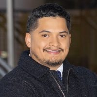 Smiling person with short dark hair and facial hair wearing a dark wool jacket over a collared shirt
