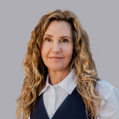 person with long, curly blond hair wearing a white button up under a gray vest.